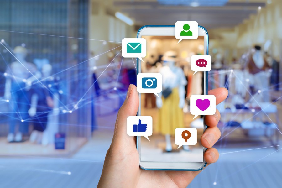 Hand holding a phone in front of a mall retail store with social media icons on the front of the phone.